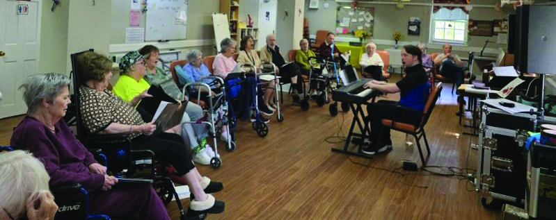 Group of people sitting in a semi circle in front a person with a keyboard piano