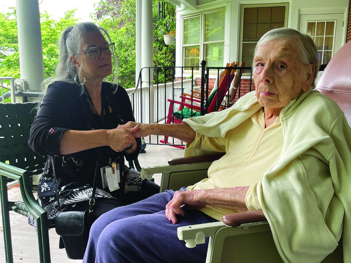 Two women sitting together on a porch, one holds the other's hand.