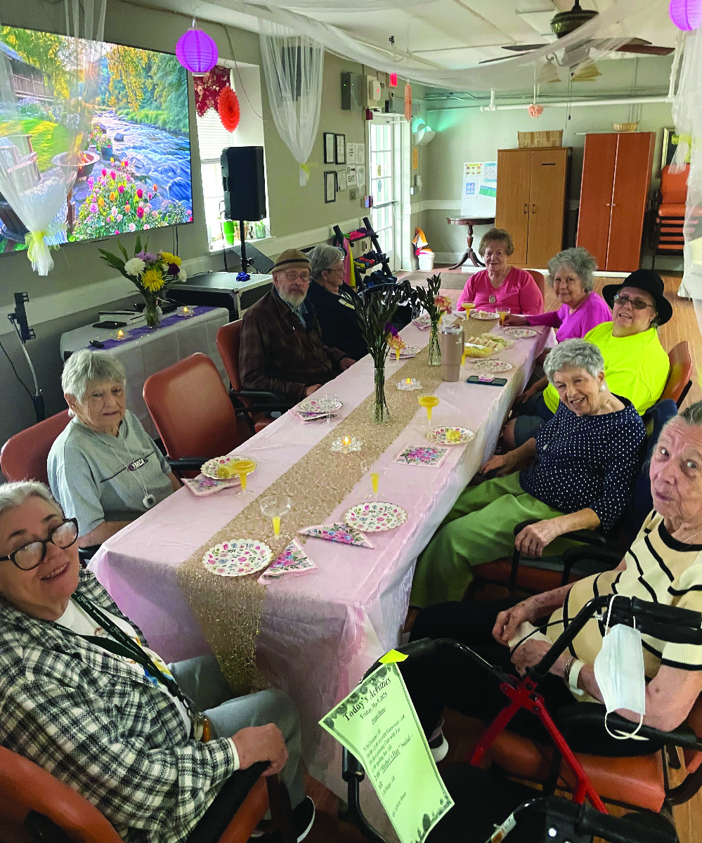 A group of people are sitting around a long rectangular table decorated with a pink tablecloth and place settings for Mother's Day
